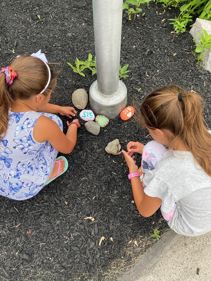Picture of two young students placing painted rocks at the bottom of a flagpole in memory of residential school children